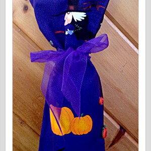 May include: A Halloween-themed gift bag in deep blue, adorned with orange pumpkins, a witch, and a crow. The bag is tied with a sheer purple ribbon. The design includes a tombstone and a witch's hat.