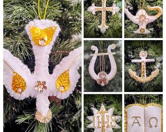 May include: A collection of handmade Christmas ornaments in various shapes, including a dove, cross, lamp, lyre, anchor, star, and book. Each ornament is white with gold accents, sequins, and beads, hanging on a Christmas tree.