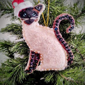 May include: A Siamese cat Christmas ornament made of felt, wearing a Santa hat. The cat has a cream-colored body, dark brown face, and a tail adorned with dark sequins. The ornament is embellished with small white beads.