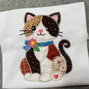 May include: A white t-shirt featuring a large, colourful appliqué of a sitting cat. The cat is constructed from various fabric patches in brown, cream, and floral patterns, with a blue collar, a pink flower, and a small pink heart on its paw.