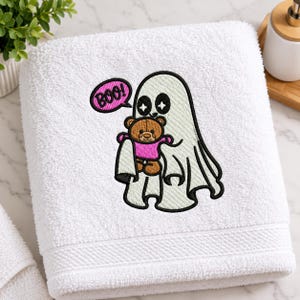 May include: A white terry cloth towel featuring an embroidered design of a white ghost with star-shaped eyes holding a brown teddy bear wearing a pink shirt. A pink speech bubble with a black outline contains the text BOO! in black letters.