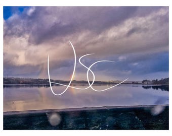 Pode incluir: Uma vista paisagística de um corpo de água calmo que reflete um céu nublado com um arco-íris ténue visível ao longe. Um logótipo em escrita branca estilizada com as letras USC está sobreposto no centro da imagem sobre o horizonte.