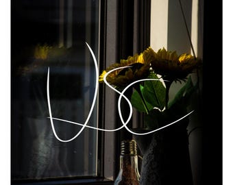Pode incluir: Um jarro decorativo com um padrão de renda contém girassóis amarelos, posicionado ao lado de uma lâmpada de vidro transparente num parapeito. A cena é iluminada pela luz solar natural, projetando sombras na moldura escura da janela.