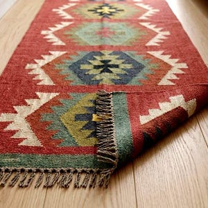 May include: A rectangular woven runner rug featuring a deep red base color with repeating geometric patterns in shades of blue, green, yellow, and white. The rug has a textured surface and is finished with short, knotted fringe along the edges.