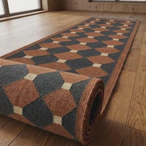 May include: A long runner rug featuring a repeating geometric pattern of dark gray and terracotta brown diamonds separated by small cream-colored squares. The rug is partially rolled at one end, revealing its textured pile and finished edges.