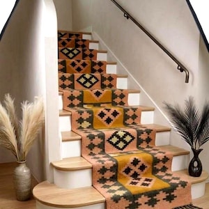 May include: A long stair runner rug featuring a repeating geometric pattern in shades of muted orange, mustard yellow, and dark charcoal gray. The runner has a textured pile and is finished with short, dark fringe at the bottom edge of the stairs.