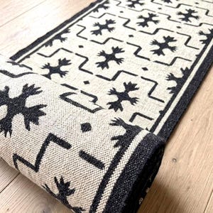 Puede incluir: Una alfombra rectangular con una base de color crema y un patrón geométrico negro repetitivo de líneas en zigzag y motivos florales estilizados. La alfombra tiene un borde negro grueso y está parcialmente enrollada.
