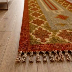 May include: A rectangular area rug featuring a geometric pattern in shades of burnt orange, olive green, and cream. The rug has a thick border with a repeating diamond motif and a central design, finished with short, knotted fringe along the edge.
