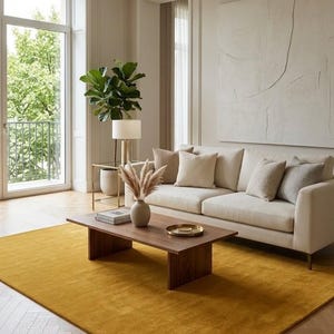 May include: A living room featuring a cream-colored sofa with four throw pillows, a rectangular wooden coffee table, and a large mustard yellow area rug. A vase with dried grasses sits on the table, and a tall green plant stands nearby.