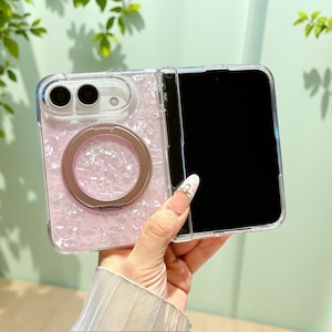 May include: A foldable smartphone case featuring a light pink pearlescent shell pattern on the back panel. The case includes a circular rose gold metal ring stand integrated into the center and a clear frame surrounding the device's camera module.