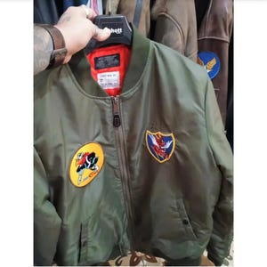 Può includere: Un giubbotto bomber verde oliva con fodera interna arancione brillante, chiusura frontale con zip e due toppe ricamate sul petto. La toppa sinistra è circolare con un uccello dei cartoni animati, quella destra è a forma di scudo con una creatura.