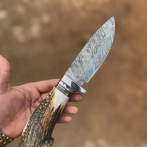 Peut inclure: Un couteau à lame fixe doté d'une lame en acier damas à motifs, présentant un design métallique ondulé et stratifié. Le manche est fabriqué en bois de cerf texturé brun et crème, rehaussé par une garde et une mitre en métal argenté poli.