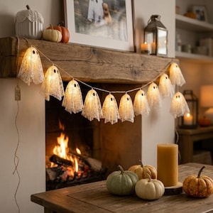 May include: A string of ten illuminated ghost-shaped lights made of white lace fabric with black felt eyes. Each ghost is connected by a thin twine cord. The ghosts are draped across a rustic wooden mantelpiece, glowing with a warm light.