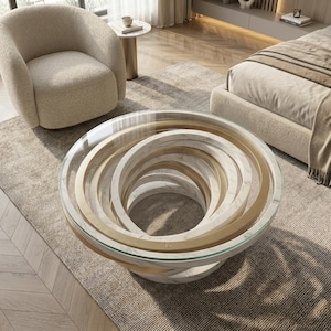 Könnte beinhalten: Ein runder Couchtisch mit einer klaren Glasplatte und einem skulpturalen Untergestell aus ineinandergreifenden kreisförmigen Ringen. Das Gestell wechselt zwischen einer weißen Marmoroptik und einem Goldfinish, was einen geschichteten Spiraleffekt erzeugt.