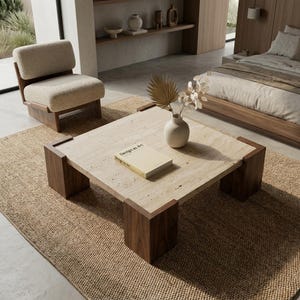 Puede incluir: Una mesa de centro cuadrada con una superficie de piedra travertino beige y cuatro patas de esquina gruesas de madera oscura. Un libro titulado Design as Art descansa sobre la superficie junto a un jarrón de cerámica con tallos secos y una hoja de palma.