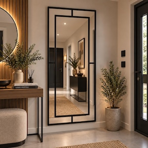 May include: A tall, rectangular wall mirror featuring a sleek black metal frame with a geometric grid design. The mirror is mounted vertically on a light-colored wall, reflecting a hallway interior with a console table, a plant, and a doorway.
