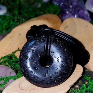 Op de afbeelding: Een zwarte lavasteen hanger met een poreus, getextureerd oppervlak in de vorm van een donut. De ronde steen hangt aan een glad, zwart leren koord dat door het middelste gat loopt, wat zorgt voor een eenvoudig en donker kettingontwerp.