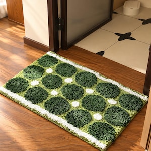May include: A rectangular bath mat featuring a tufted, shaggy texture with a repeating pattern of dark green circles on a light green background, accented by small white circles and white border stripes along the edges.
