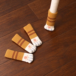 May include: Four knitted chair leg covers featuring a mustard yellow and brown striped pattern. Each cover has a white base with a pink paw pad design and light blue toe accents. One cover is fitted onto a white chair leg, while three lie flat.