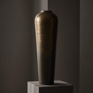 May include: A tall, cylindrical vase with a tapered base and a narrow opening. The upper portion features a textured, metallic gold speckled finish that transitions into a solid, dark matte brown color toward the bottom of the vessel.
