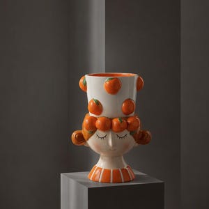 Pode incluir: Um vaso de cerâmica em forma de cabeça com base branca e detalhes de frutas laranja. A figura tem olhos fechados, um pequeno sorriso e cabelo adornado com laranjas esculpidas. A parte superior é um recipiente cilíndrico.