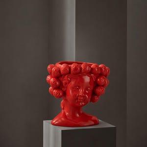 Pode incluir: Um vaso de cerâmica vermelho brilhante em forma de cabeça humana, com acabamento lustroso e uma coroa decorativa de elementos texturizados em forma de fruta esférica que circundam a abertura superior do recipiente.