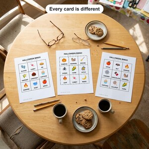 May include: Three white paper Halloween Bingo game cards with colorful illustrations and text labels, two pairs of eyeglasses, two pencils, and two plates with chocolate chip cookies, all arranged on a round wooden table.
