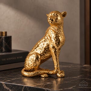 Könnte beinhalten: Eine goldfarbene Leopardenfigur in sitzender Position mit einer texturierten, gefleckten Oberfläche. Die Skulptur zeigt einen detaillierten Kopf, aufmerksame Ohren und einen langen Schwanz, der um die Basis gewickelt ist, mit glänzendem Finish.