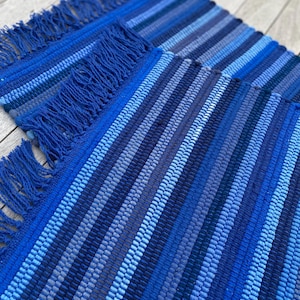May include: Two blue woven placemats with a striped pattern in various shades of blue. Each placemat has a fringe along one edge. The placemats are made of a textured material and are suitable for dining.