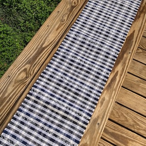 May include: A rectangular table runner with a blue, white, and black checkered pattern. The runner is made of woven fabric and is displayed on a wooden deck. The runner is a decorative item for a table.