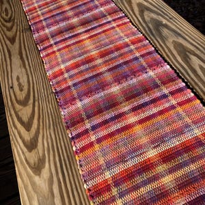 May include: A handwoven table runner with a vibrant, striped pattern in shades of red, orange, purple, and yellow. The runner is made of woven fabric and is displayed on a wooden surface.