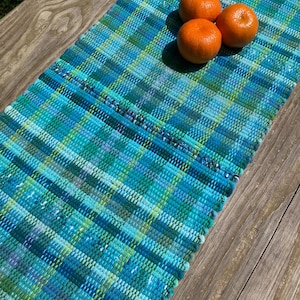 May include: A handwoven table runner with a plaid pattern in shades of blue and green. Three oranges rest on the runner. The runner is made of a textured fabric and is displayed on a wooden surface.