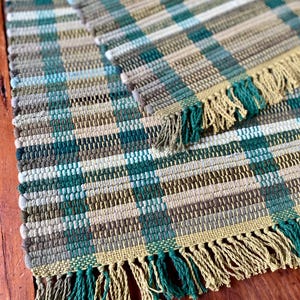 May include: Handwoven rectangular rug with a plaid pattern in shades of teal, green, beige, and brown. The rug has a fringed edge in teal and beige. The rug is made of a woven material.