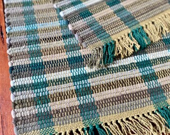 May include: Handwoven rectangular rug with a plaid pattern in shades of teal, green, beige, and brown. The rug has a fringed edge in teal and beige. The rug is made of a woven material.
