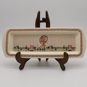 May include: A rectangular cream-coloured ceramic tray featuring a decorative border of small red flowers. The centre depicts a landscape with green trees, small houses, and a figure holding a red umbrella, all set against a vertical striped background.