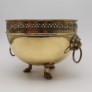 May include: A polished brass bowl featuring a decorative openwork gallery rim, three clawed feet, and two side handles shaped like lion heads with attached rings. The metal surface is smooth and reflective with a warm golden metallic tone.