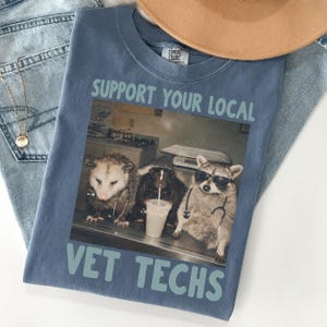 May include: A blue t-shirt featuring a graphic print of an opossum, a skunk, and a raccoon wearing sunglasses and stethoscopes. The text above reads SUPPORT YOUR LOCAL and the text below reads VET TECHS. The shirt has a Comfort Colors brand label.