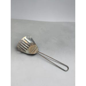 May include: A silver-colored metal kitchen spatula featuring a wide, flat head with five vertical slots and a thin, looped wire handle. The surface of the metal head shows signs of wear and discoloration, with a smooth, rounded edge at the top.