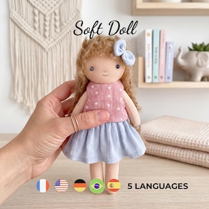 May include: A soft doll with curly brown hair, a light blue hair bow, and dark eyes. The doll wears a pink sleeveless top with white polka dots and a solid light blue skirt. The text Soft Doll is visible at the top, and 5 LANGUAGES is at the bottom.