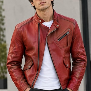 Può includere: Una giacca in pelle rossa con colletto alla coreana e bottone a pressione, una tasca diagonale con zip sul petto, due tasche inferiori con patta e chiusura a pressione, e dettagli a coste sulle maniche, indossata sopra una t-shirt bianca.