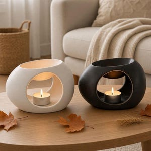 May include: Two oval-shaped ceramic oil burners, one matte white and one matte black, each featuring a central circular opening that holds a small lit tea light candle. The smooth, rounded structures provide a stable base for the candles.