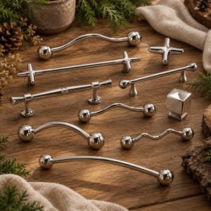 May include: A collection of polished silver-toned metal cabinet hardware, including various curved, straight, and cross-shaped drawer pulls and a single square knob, all featuring a smooth, reflective finish arranged on a wooden surface.
