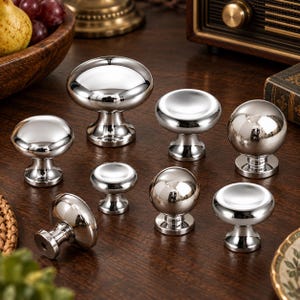 Puede incluir: Una colección de ocho pomos de metal plateado pulido para armarios en varias formas, incluyendo diseños esféricos y circulares aplanados. Cada pomo presenta una superficie lisa y reflectante con una base sólida sobre madera oscura.