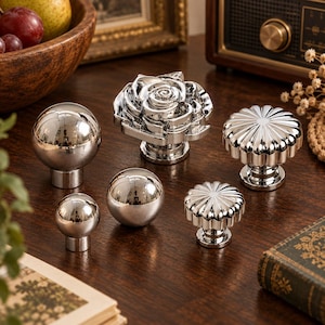 May include: A collection of six silver-toned metal cabinet knobs in various shapes, including two smooth spheres, two fluted circular designs, and one detailed rose-shaped knob, all featuring a polished, reflective finish on their surfaces.