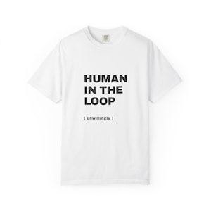 May include: A white short-sleeve t-shirt featuring the black text "HUMAN IN THE LOOP" in a bold, sans-serif font, followed by the smaller text "( unwillingly )" centered on the chest. The shirt has a standard crew neckline.