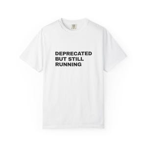 May include: A white short-sleeved t-shirt featuring the black text "DEPRECATED BUT STILL RUNNING" printed in a bold, sans-serif font across the centre of the chest. The garment has a classic crew neckline and a standard, relaxed fit design.