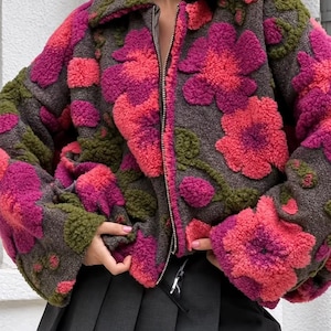 May include: A zip-up jacket made of textured, fuzzy material in a dark gray base color. The jacket features a prominent floral pattern in shades of bright pink, magenta, and olive green. It has a wide collar and long sleeves.