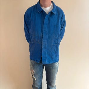 May include: A vibrant blue long-sleeve work jacket featuring a classic collar, a button-front closure, and three large patch pockets on the front. The jacket is worn over a plain white t-shirt and paired with distressed blue denim jeans.
