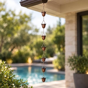 May include: A copper-coloured metal rain chain featuring a series of seven tulip-shaped cups connected by thin metal links. The chain hangs vertically from a gutter hook, with each cup designed to channel water downwards in a cascading sequence.