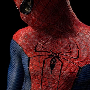 May include: A full-body suit featuring a textured red and blue fabric with a honeycomb pattern. The chest displays a large black spider emblem, and the headpiece includes dark, reflective eye lenses against a solid black background.